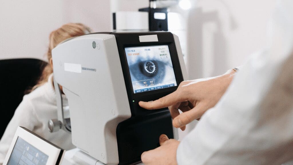 A person operates a digital eye examination machine, with another individual sitting in the background during an eye test.